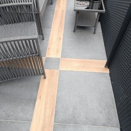 Chrystian śipek - Wykończony taras z szarymi płytkami i drewnopodobnymi wstawkami, na którym stoją szare meble rattanowe oraz stolik z kwiatami w metalowej osłonce.