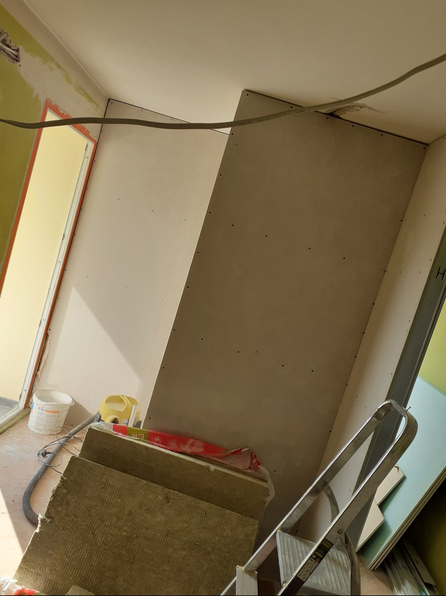 Płyty gipsowo-kartonowe zamontowane na ścianie w trakcie remontu, widoczne narzędzia i materiały budowlane: drabina, odkurzacz, wełna mineralna, wiadro z zaprawą.