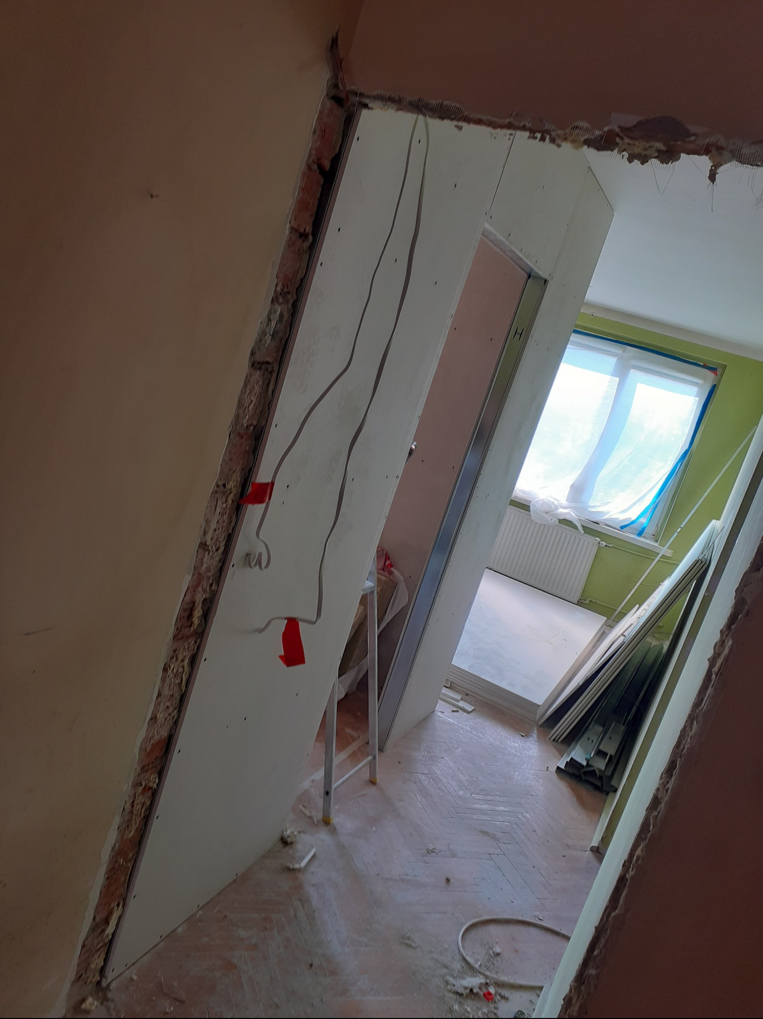 Montaż płyt kartonowo-gipsowych w trakcie remontu, widoczne kable elektryczne przytwierdzone czerwoną taśmą, surowa cegła w narożniku, podłoga z parkietu w jodełkę.