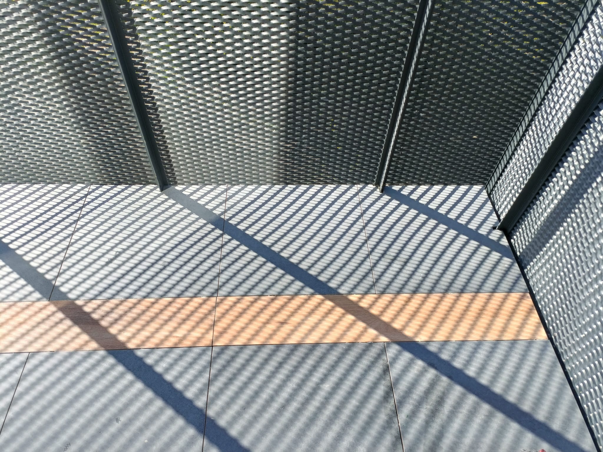 Ażurowa konstrukcja z metalu rzuca geometryczne cienie na szare płytki podłogowe z wstawką w kolorze drewna.