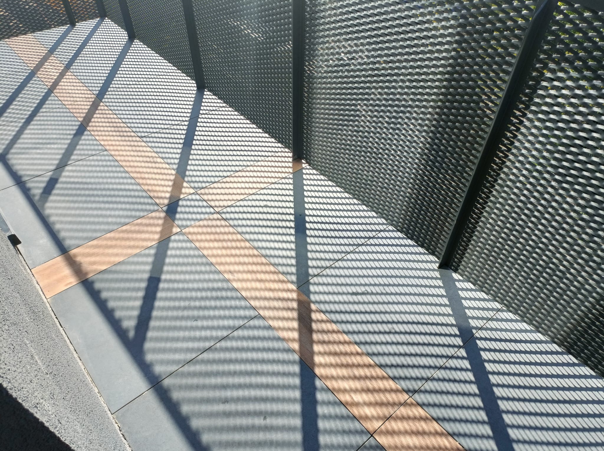 Ułożona podłoga z szarych płytek z dekoracyjnym pasem w odcieniach brązu, na którą padają cienie z metalowej ażurowej konstrukcji.