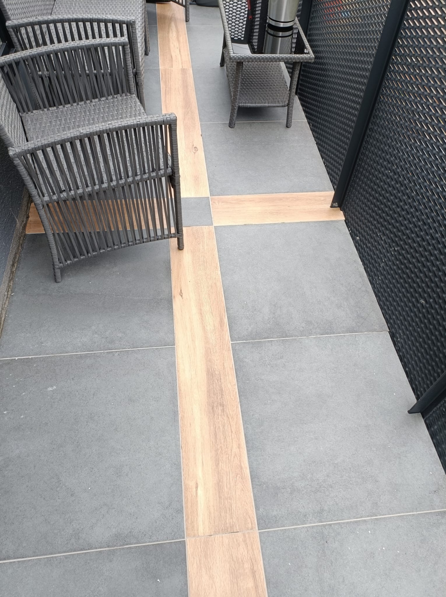 Szare płytki podłogowe na zewnątrz z drewnianymi wstawkami, uzupełnione szarymi wiklinowymi krzesłami i stolikiem, przylegające do czarnej metalowej ażurowej ściany.