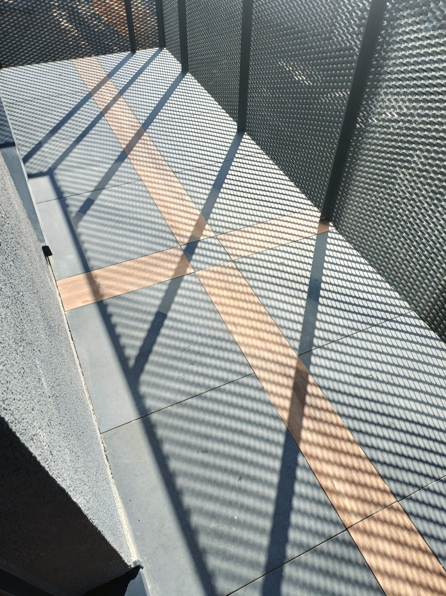 Szare płytki podłogowe z dekoracyjnymi wstawkami w odcieniu drewna, oświetlone geometrycznym cieniem metalowej konstrukcji.