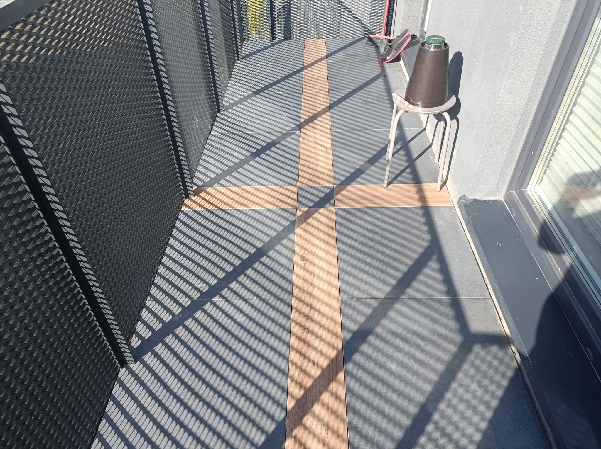 Balkon z ciemnoszarą ażurową balustradą, szarymi płytkami i wstawkami z paneli imitujących drewno, oświetlony promieniami słońca tworzącymi geometryczne cienie.