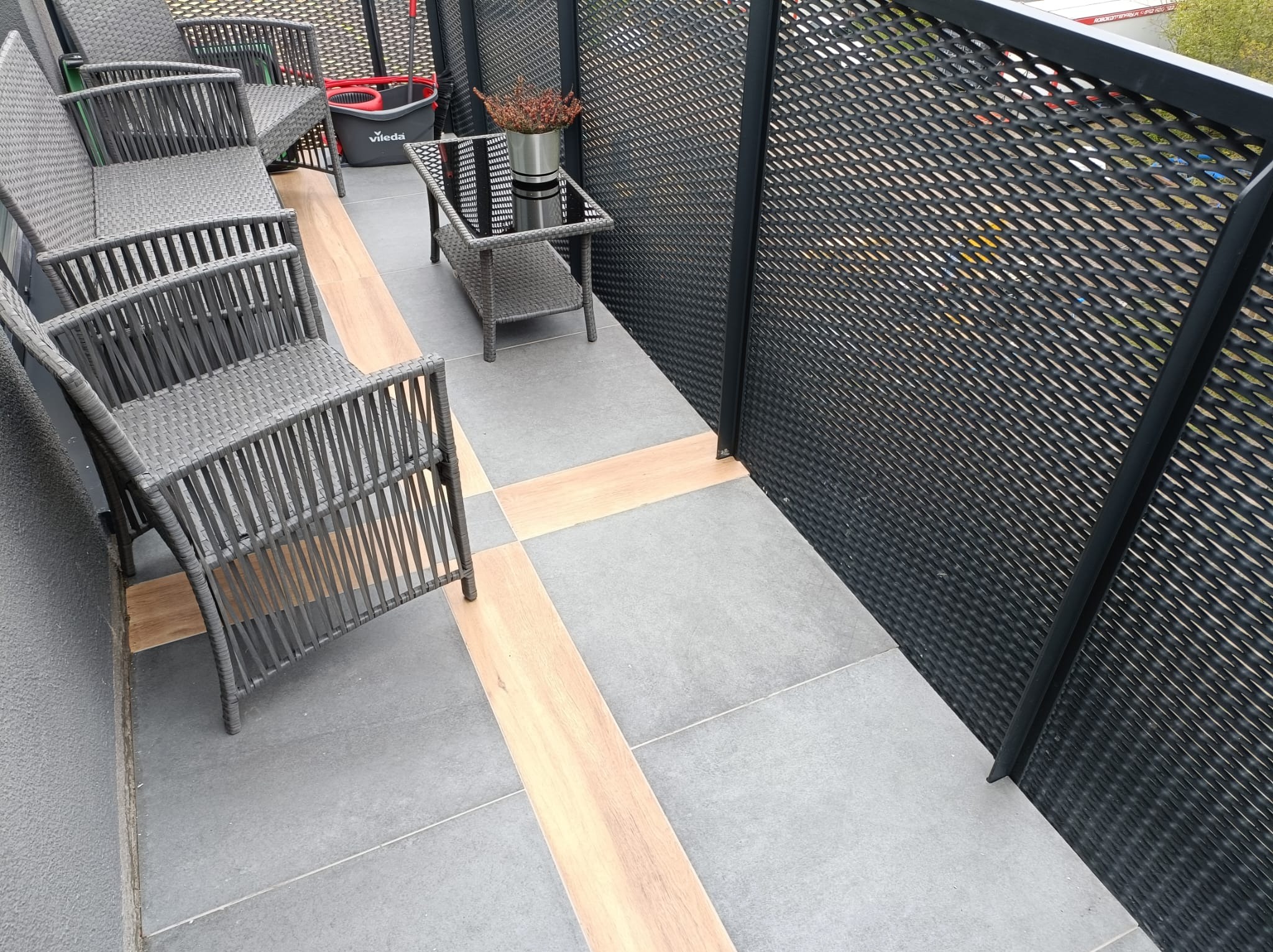 Wykończony balkon z szarymi płytkami, drewnopodobnymi wstawkami, meblami ogrodowymi z technorattanu i ażurową balustradą.