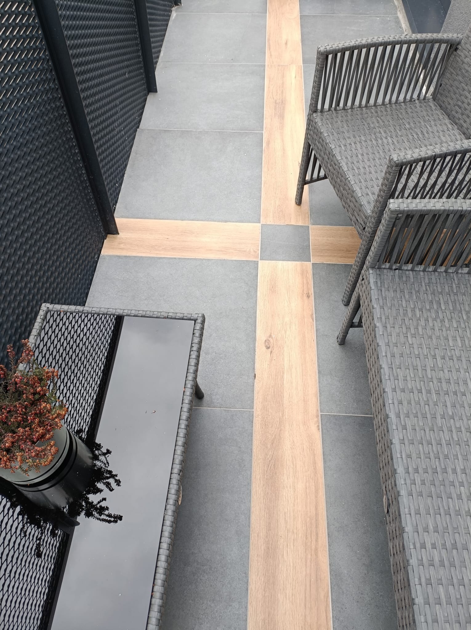 Szare płytki podłogowe z drewnopodobnymi wstawkami tworzącymi krzyż, szare meble ogrodowe z rattanu i czarny stolik z blatem z połyskliwego szkła, na którym stoi doniczka z brązowymi kwiatami.
