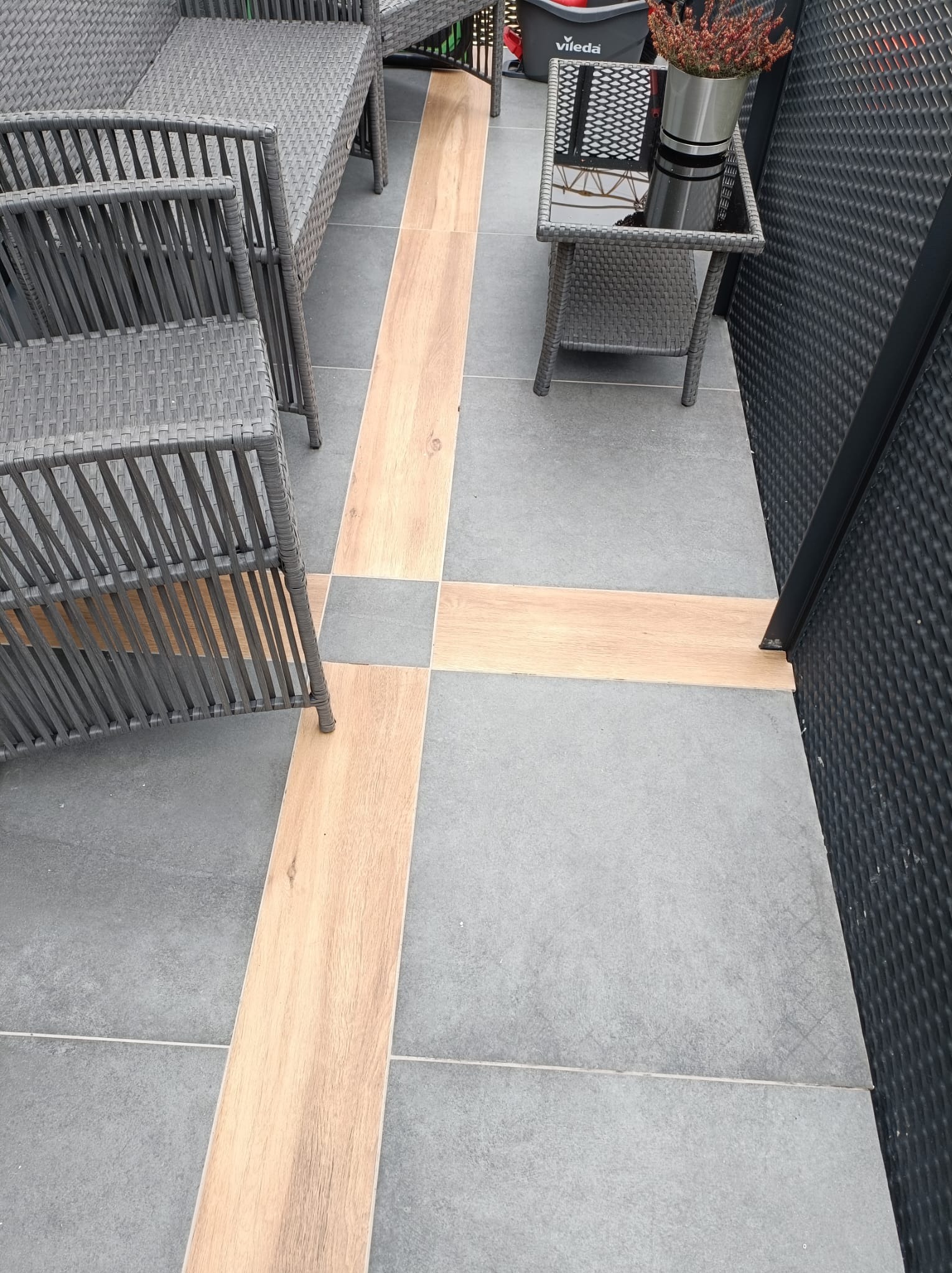 Wykończony taras z szarymi płytkami i drewnopodobnymi wstawkami, na którym stoją szare meble rattanowe oraz stolik z kwiatami w metalowej osłonce.