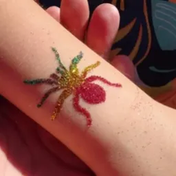 Błyszczący, kolorowy tatuaż brokatowy w kształcie pająka na ramieniu dziecka, wykonany z czerwonego, złotego i zielonego brokatu.