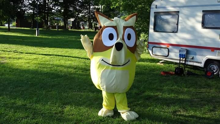 Osoba w kostiumie psa na zielonym trawniku, z przyczepą kempingową w tle.