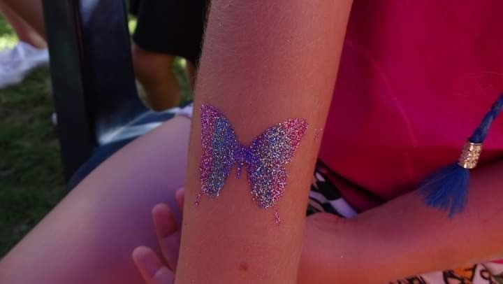 Brokatowy tatuaż w kształcie motyla na ramieniu dziecka, z widoczną niebieską ozdobą na ubraniu.
