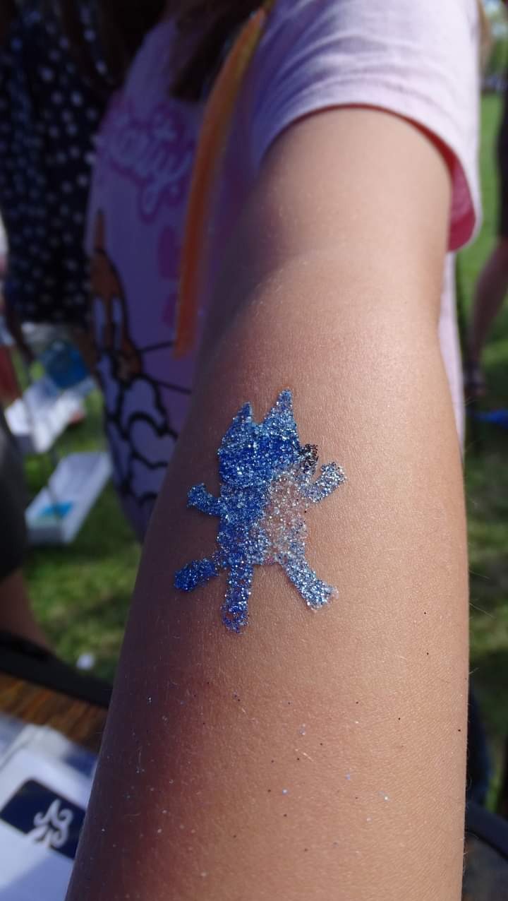 Błyszczący, niebieski tatuaż brokatowy w kształcie postaci z bajki na ramieniu dziecka, wykonany podczas imprezy plenerowej.