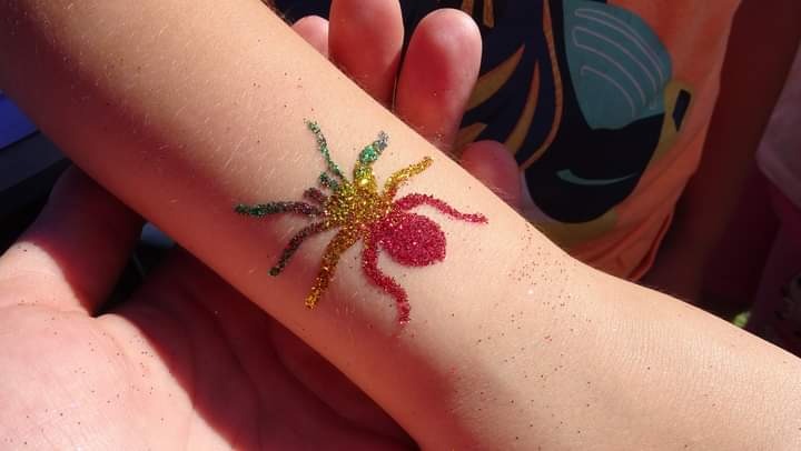 Błyszczący, kolorowy tatuaż brokatowy w kształcie pająka na ramieniu dziecka, wykonany z czerwonego, złotego i zielonego brokatu.