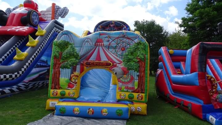 Zestaw dmuchańców na trawie, w tym dmuchany zamek z motywem cyrku i dmuchana zjeżdżalnia z motywem wyścigów samochodowych.