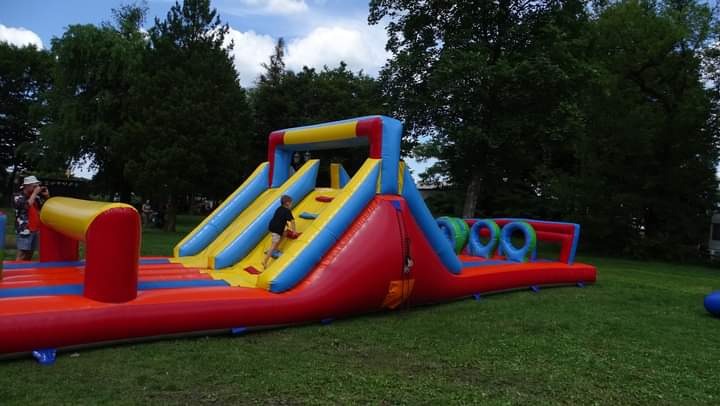 Dmuchany tor przeszkód z dwoma zjeżdżalniami, drabinką i tunelem z oponami, na trawie, z dzieckiem wspinającym się na drabinkę, w tle drzewa i osoby.