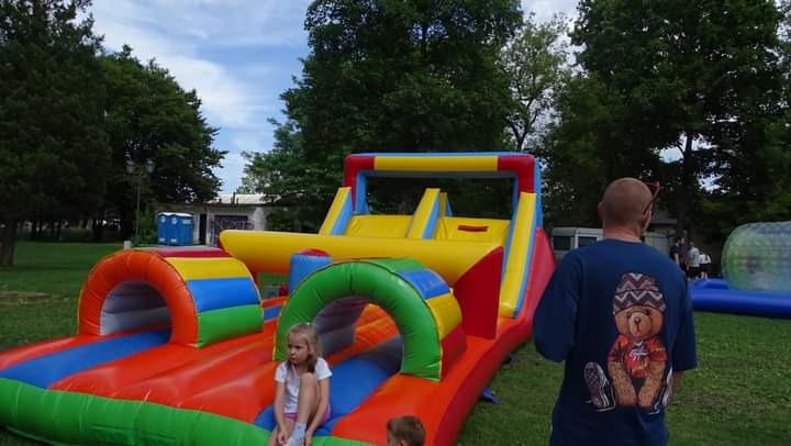 Kolorowy tor przeszkód dmuchany na trawie, z dziećmi i dorosłym w tle, częściowo widoczna dmuchana kula.