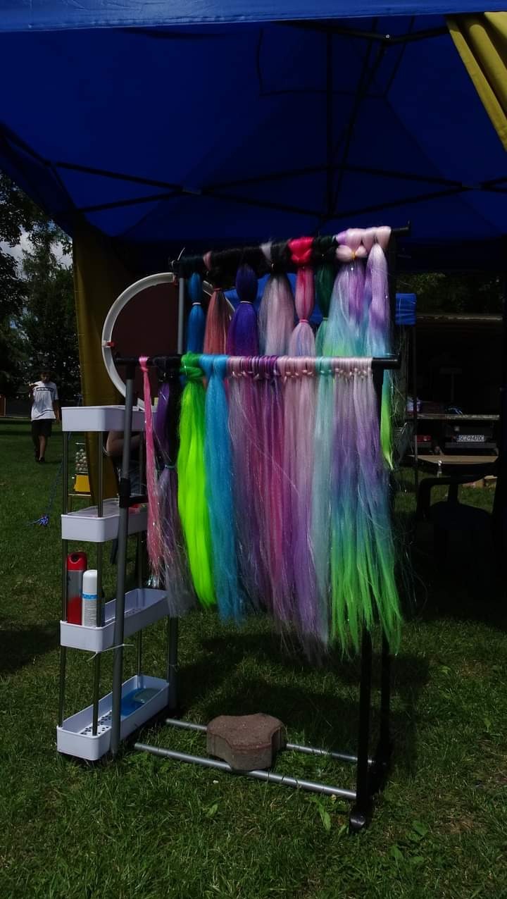 Stoisko plenerowe z kolorowymi pasmami włosów syntetycznych na metalowym wieszaku, widoczne kosmetyki na półce, trawa i fragment namiotu w tle.