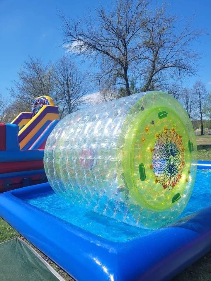 Napełniona wodą, przezroczysta, dmuchana kula z zielonymi elementami na obwodzie, umieszczona w niebieskim basenie, w tle dmuchana zjeżdżalnia i drzewa bez liści.