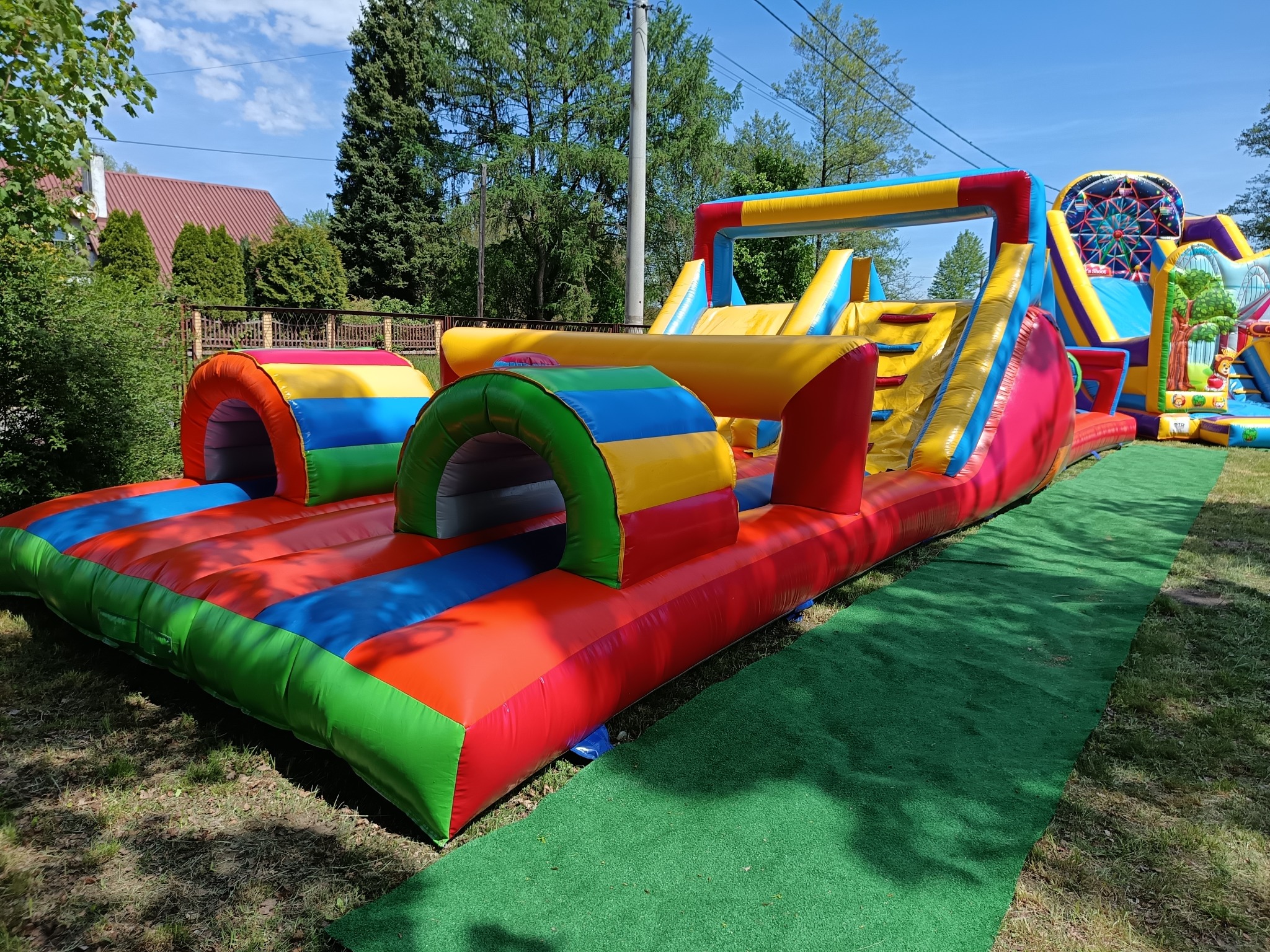 Kolorowy, rozłożysty dmuchany plac zabaw z tunelem i zjeżdżalnią, rozstawiony na trawie w ogrodzie. Widoczny fragment domu w tle, błękitne niebo.