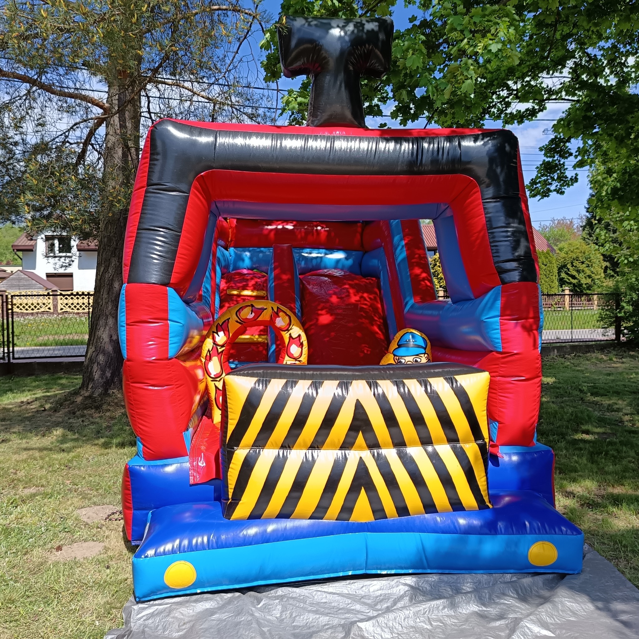 Kolorowy, dmuchany zamek w kształcie wozu strażackiego, ustawiony na trawie w ogrodzie. Widoczne elementy graficzne i zjeżdżalnia wewnątrz.