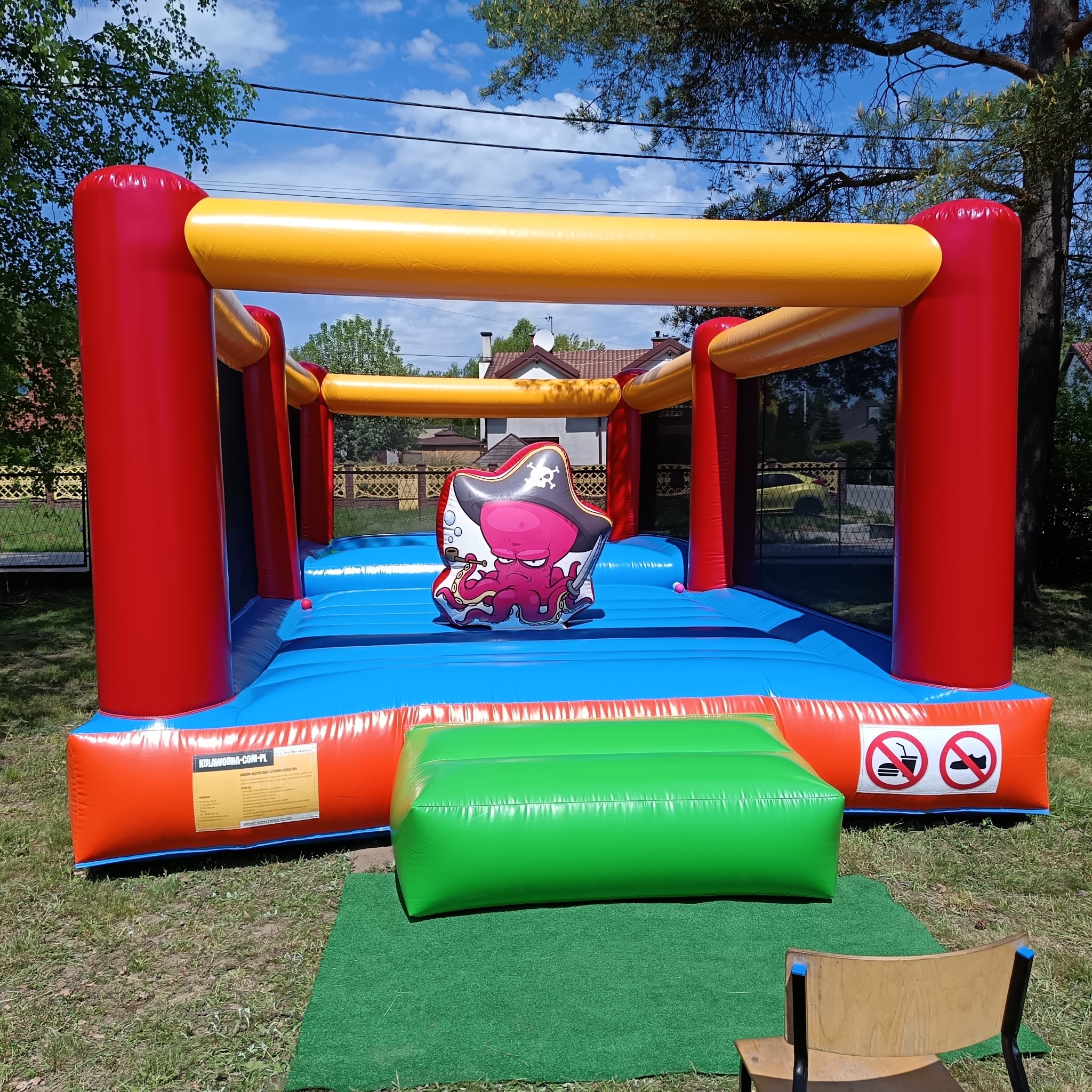 Kolorowy dmuchany zamek z motywem pirackiego ośmiornicy na trawie, z widocznymi znakami zakazu jedzenia i zdejmowania obuwia. Słoneczny dzień.