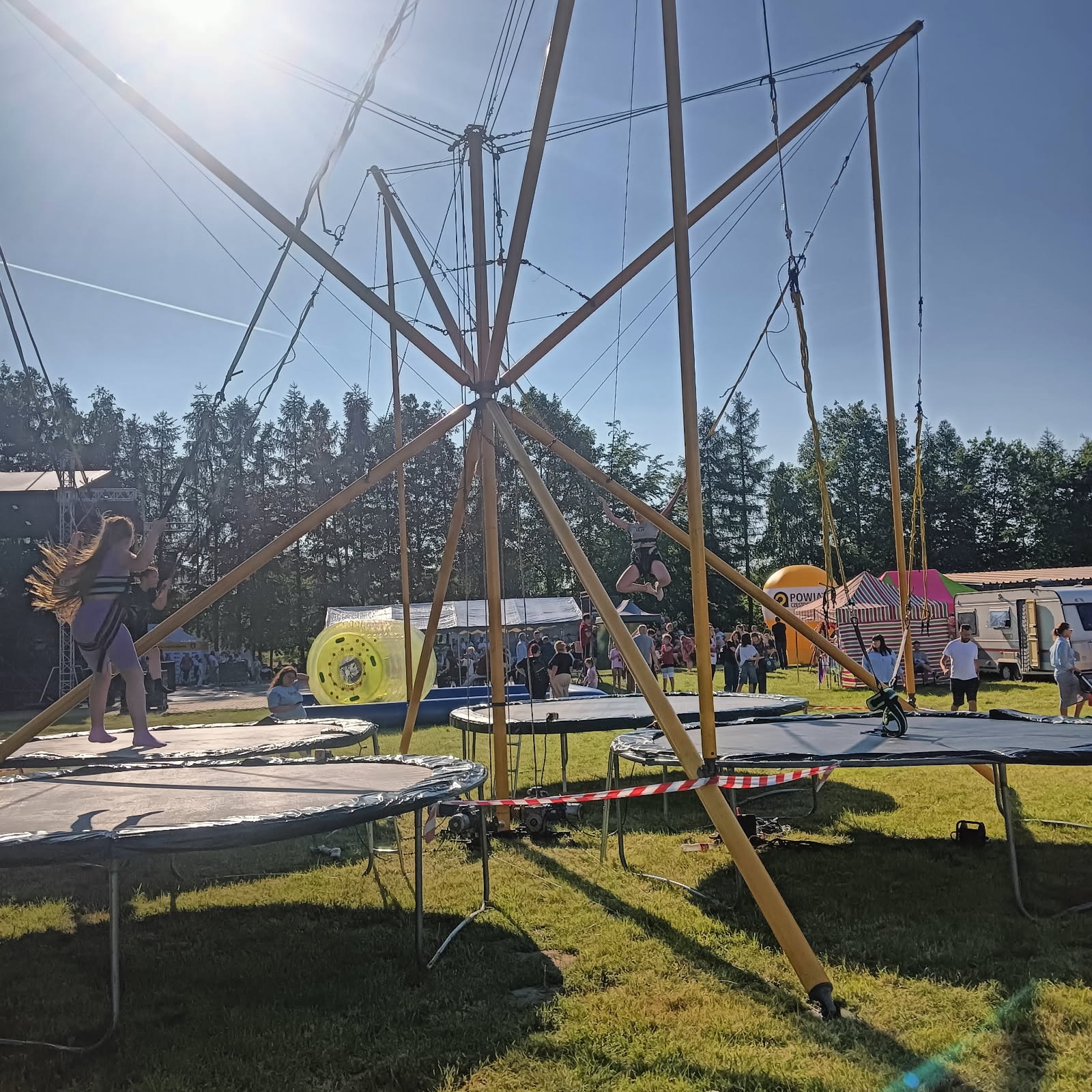 Park trampolin z konstrukcją do skoków na gumie. Dzieci skaczą na trampolinach, w tle tłum ludzi, drzewa i dmuchany walec. Słoneczny dzień.