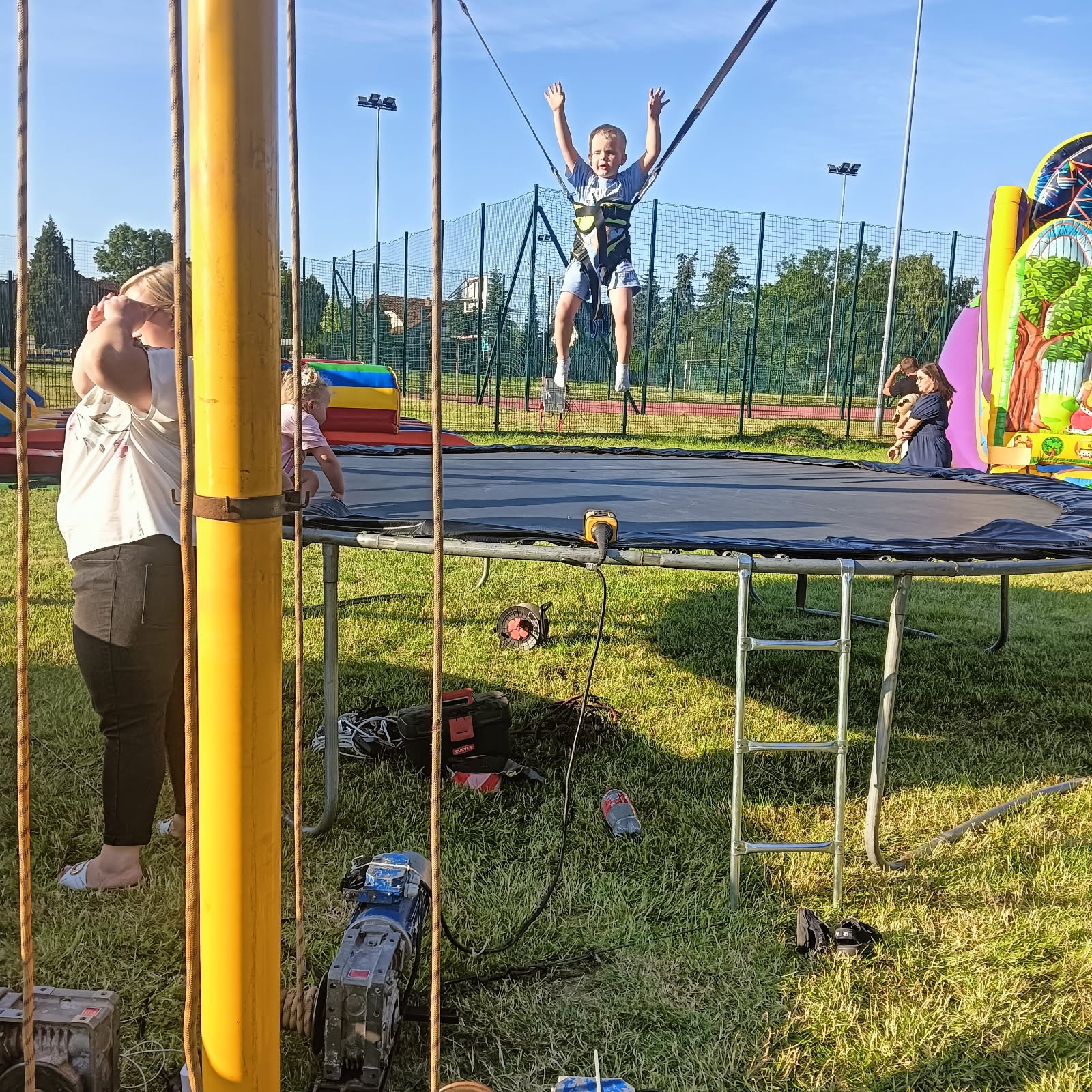 Chłopiec skacze na trampolinie bungee z uprzężą, w tle dmuchany zamek i publiczność. Zabawa na świeżym powietrzu podczas imprezy plenerowej.