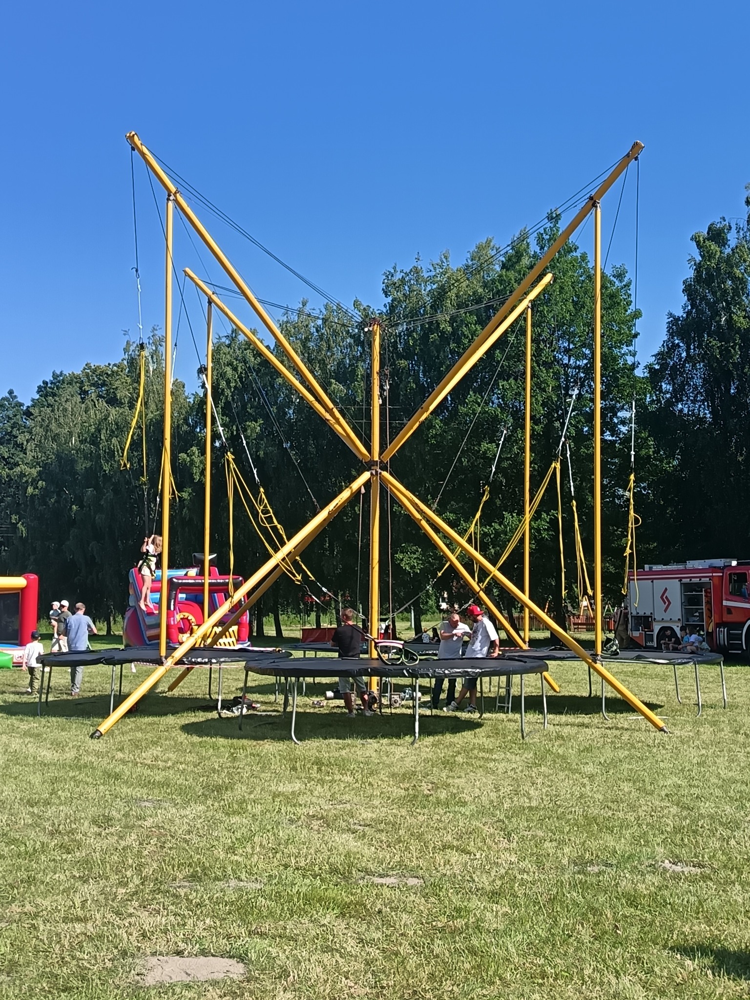 Żółta konstrukcja trampoliny bungee na zielonej trawie, dzieci korzystające z atrakcji. W tle dmuchany zamek i wóz strażacki, błękitne niebo.