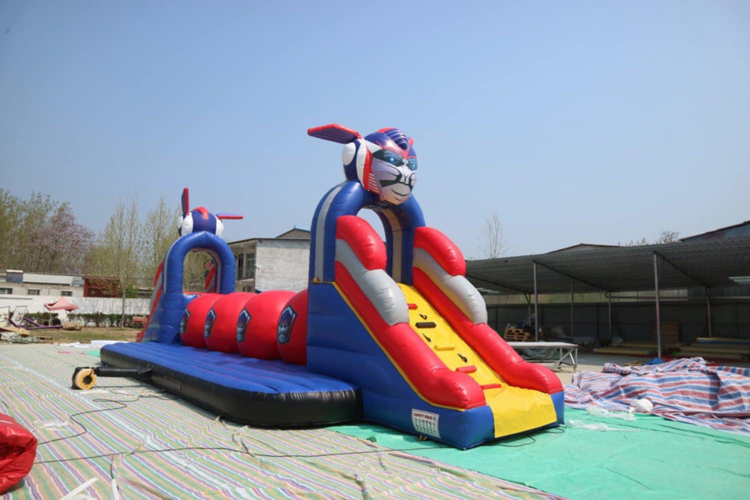 Kolorowy dmuchany zamek z tunelem i zjeżdżalnią w kształcie robota, ustawiony na zewnątrz, na zielonej plandece. Widoczne budynki w tle.