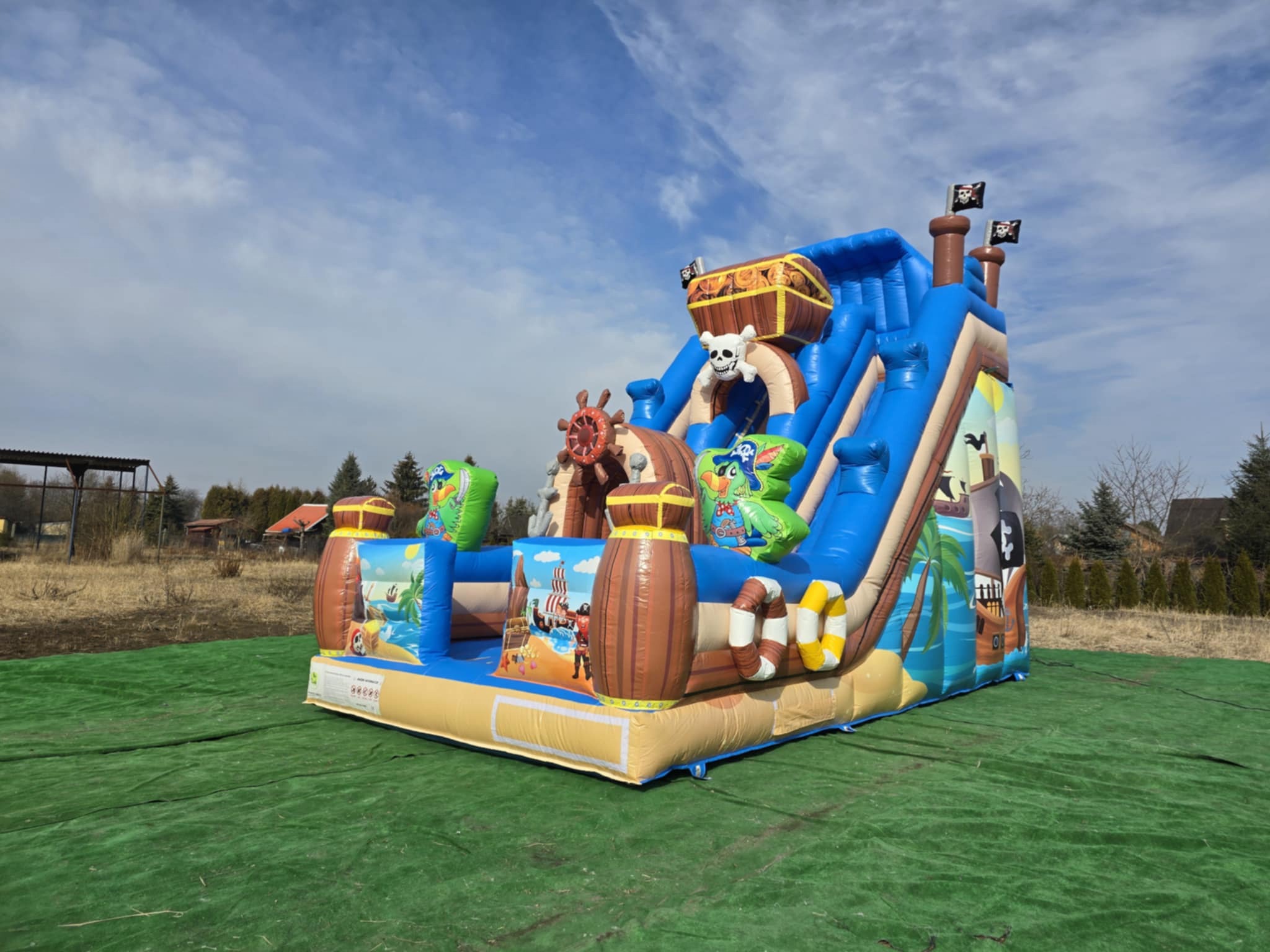 Nadmuchiwany zamek w pirackim stylu z motywami skrzyń, beczek i żeglugi, ustawiony na zielonej macie na zewnątrz, pod błękitnym niebem.