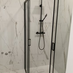 Mariusz Zaniewicz - Nowoczesna kabina prysznicowa z czarnymi profilami i deszczownicą na tle marmurowych płytek w odcieniach bieli i szarości. Minimalistyczny design, widoczne detale wykończenia.