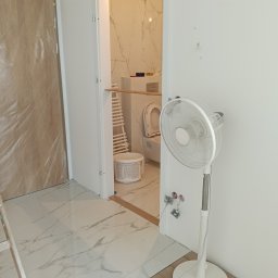 Mariusz Zaniewicz - Wykończona łazienka z marmurowymi płytkami widoczna przez otwarte drzwi. Podłoga wyłożona panelami, biały wentylator stojący, drabina i wiadro z farbą w tle.