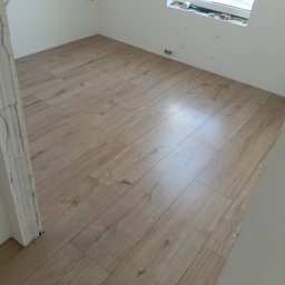 Mariusz Zaniewicz - Jasne panele podłogowe ułożone w pokoju z białymi ścianami i oknem. Widoczne niedokończone wykończenia przy ścianach i futrynie.