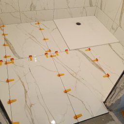 Mariusz Zaniewicz - Białe płytki imitujące marmur, ułożone w łazience, z pomarańczowymi klipsami poziomującymi. Widoczny odpływ liniowy i fragment ściany z otworami na armaturę.