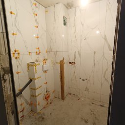 Mariusz Zaniewicz - Białe płytki imitujące marmur na ścianach łazienki w trakcie montażu, z użyciem pomarańczowych krzyżyków dystansowych i taśmy maskującej na stelażu podtynkowym WC.