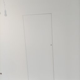 Mariusz Zaniewicz - Biała, ukryta w ścianie, minimalistyczna klapa rewizyjna z małym, okrągłym zamkiem, w świeżo pomalowanym pomieszczeniu. Widoczne ślady po remoncie na podłodze.