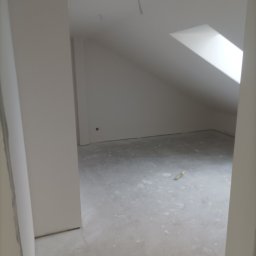 Mariusz Zaniewicz - Poddasze ze skośnym oknem dachowym w trakcie prac wykończeniowych. Widoczna szpachla na ścianach i podłodze, przygotowanie do dalszych prac. Minimalistyczny kadr.