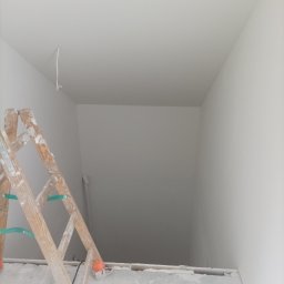 Mariusz Zaniewicz - Świeżo pomalowane na biało wnętrze z drewnianą drabiną malarską. Widoczny kabel wystający z sufitu. Minimalistyczne ujęcie po malowaniu.