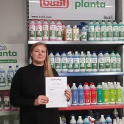 Kobieta trzymająca certyfikat na tle regału z produktami do sprzątania marki Buzil Planta, prezentacja uczestnictwa w szkoleniu.