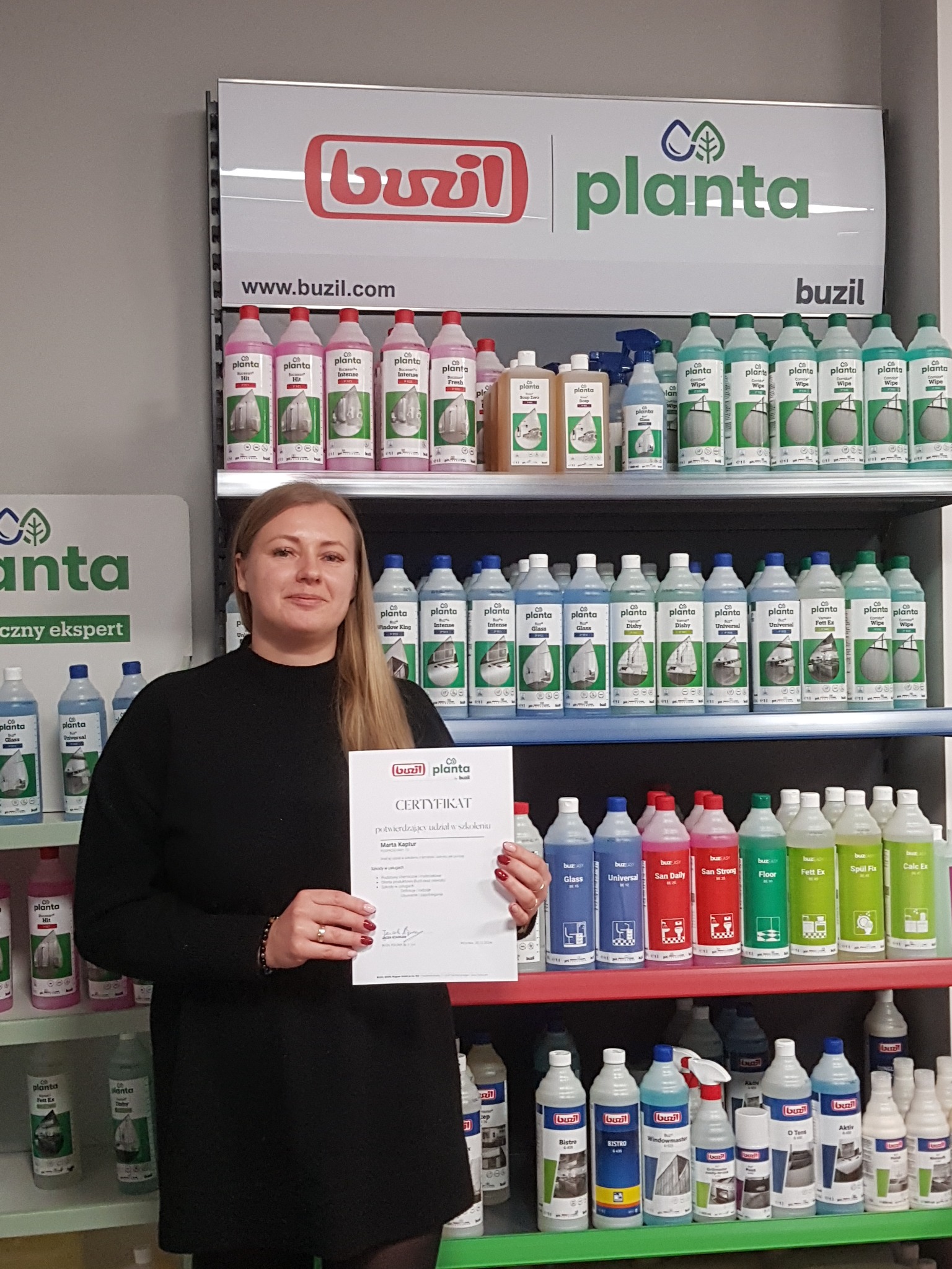 Kobieta trzymająca certyfikat na tle regału z produktami do sprzątania marki Buzil Planta, prezentacja uczestnictwa w szkoleniu.