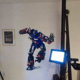 Adam Marcinkowski DPI DRUK ŚCIENNY POZNAŃ - Robot Transformer w niebiesko-czerwonych barwach nanoszony na ścianę za pomocą pionowej drukarki, obok wisi obrazek z napisem Dream BIG!