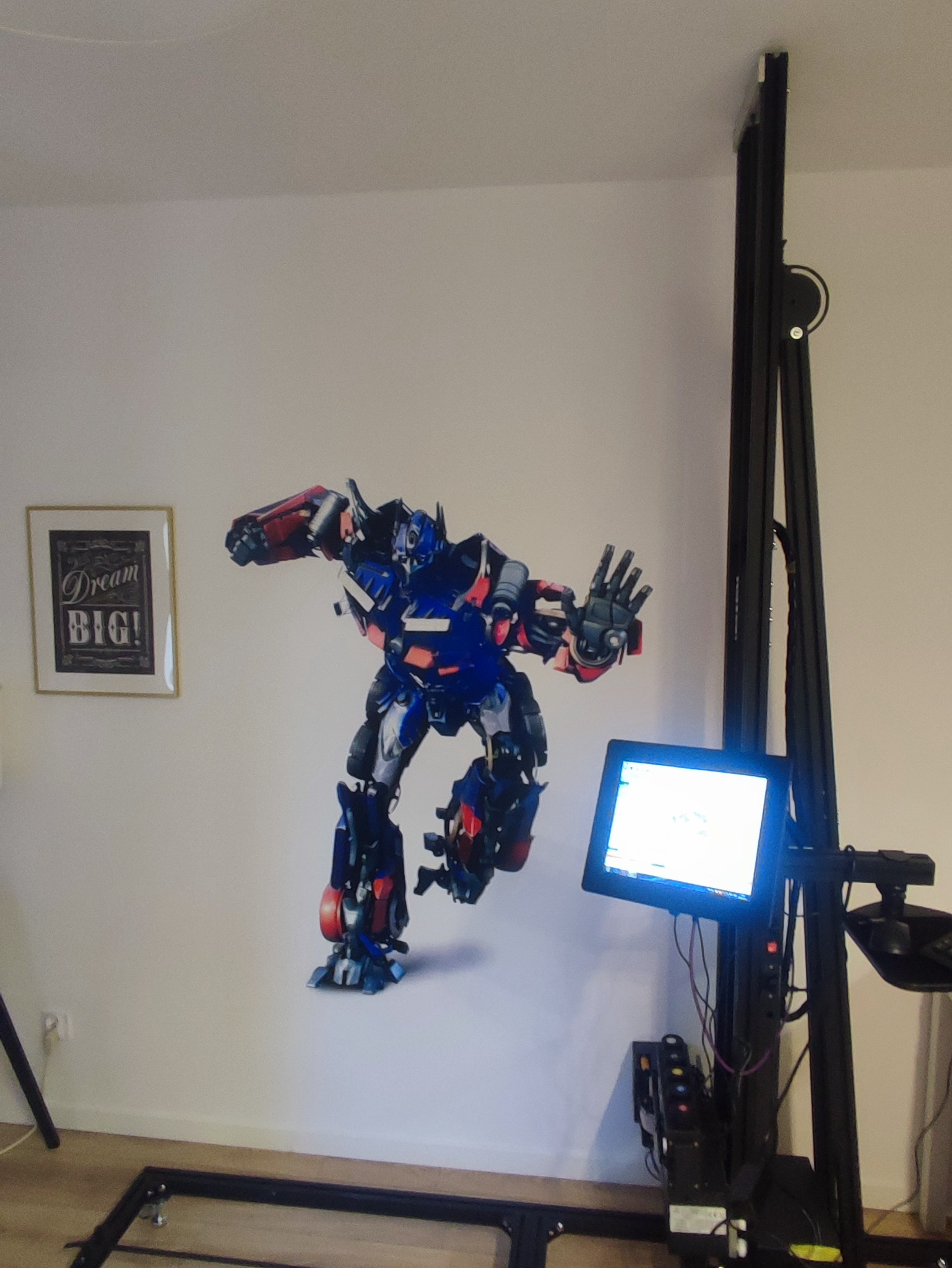 Robot Transformer w niebiesko-czerwonych barwach nanoszony na ścianę za pomocą pionowej drukarki, obok wisi obrazek z napisem Dream BIG!