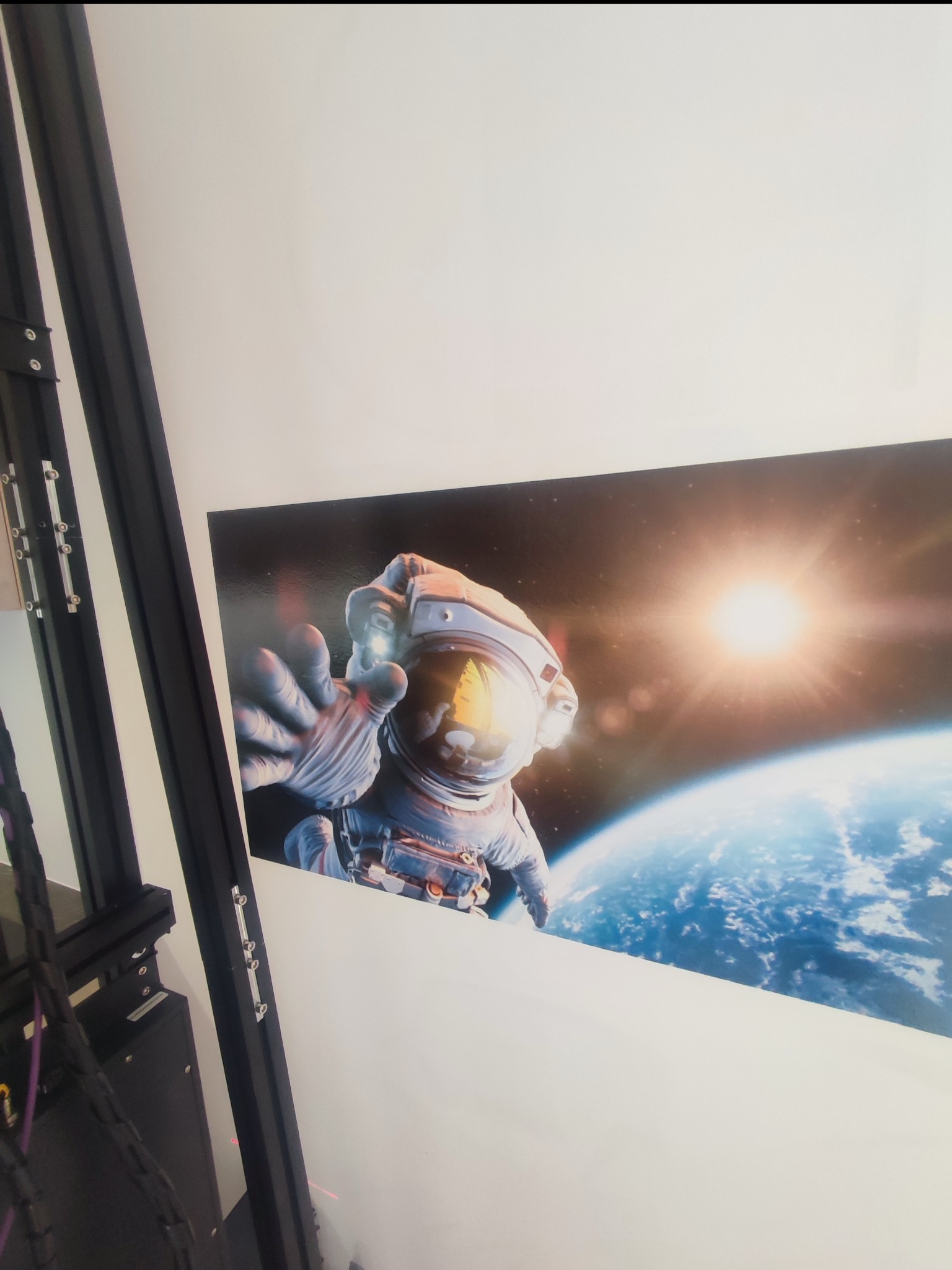 Zdjęcie przedstawia wydruk plakatu z motywem astronauty na tle Ziemi i kosmosu, zawieszonego na białej ścianie w biurze, widoczne elementy ramy drukarki 3D w lewym dolnym rogu.