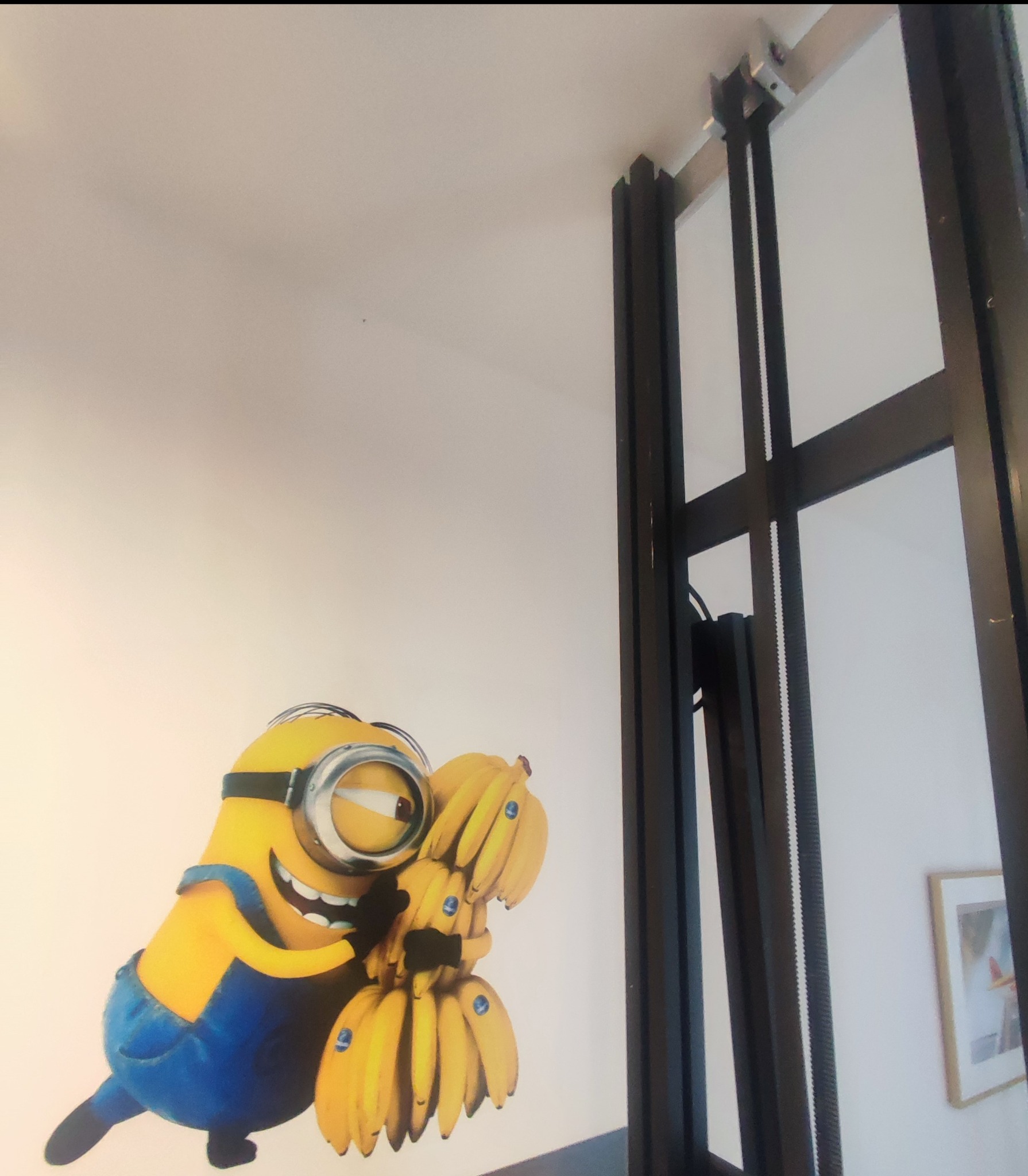 Naklejka na ścianie w biurze przedstawiająca Minionka przytulającego kiść bananów, obok czarnej ramy drzwi.