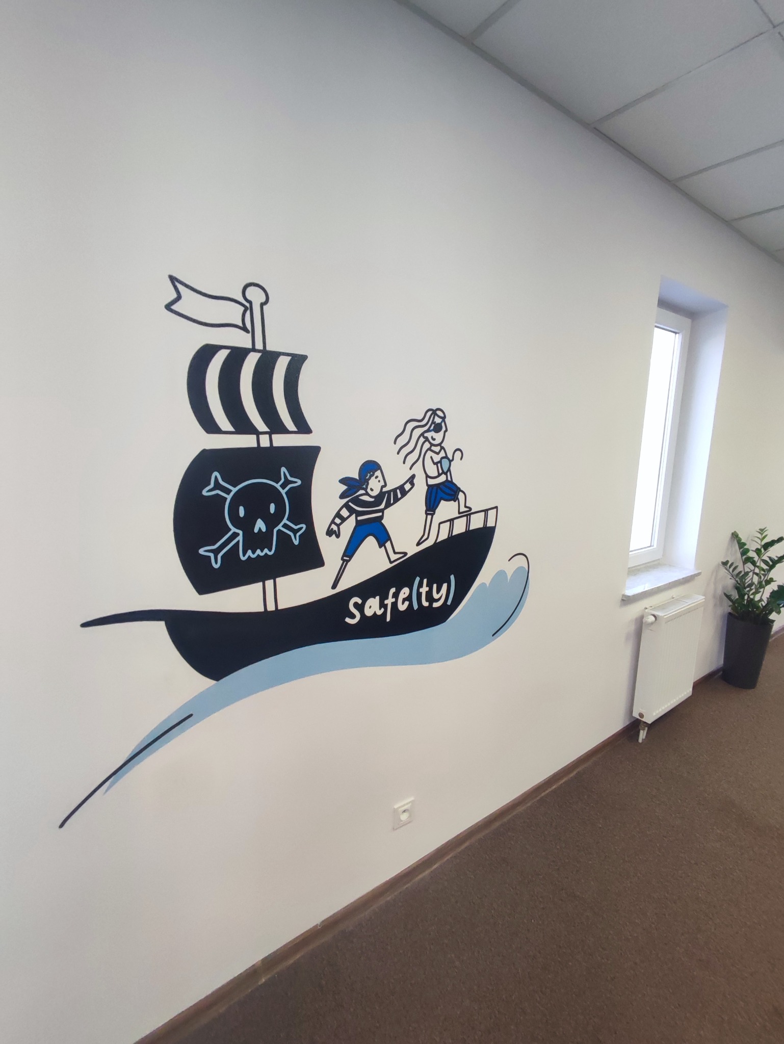 Biała ściana biura z ręcznie malowaną ilustracją pirackiego statku z napisem 'Safe(ty)', obok okno i roślina doniczkowa.