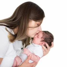 Matka w białej koszulce delikatnie całuje w czoło śpiącego noworodka ubranego w białe body na jasnym tle.