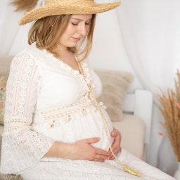FOTO MAGIA Paulina Kryszczyńska - Kobieta w ciąży w białej koronkowej sukience i słomkowym kapeluszu, siedząca na puszystym dywanie, trzyma ręce na brzuchu, patrząc w dół.