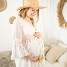 FOTO MAGIA Paulina Kryszczyńska - Kobieta w ciąży w białej koronkowej sukience i słomkowym kapeluszu, trzymająca ręce na brzuchu, w tle dekoracje boho z suszonych traw.