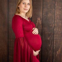 FOTO MAGIA Paulina Kryszczyńska - Kobieta w czerwonej sukni ciążowej z rękawami z szyfonu pozuje na tle drewnianej ściany.