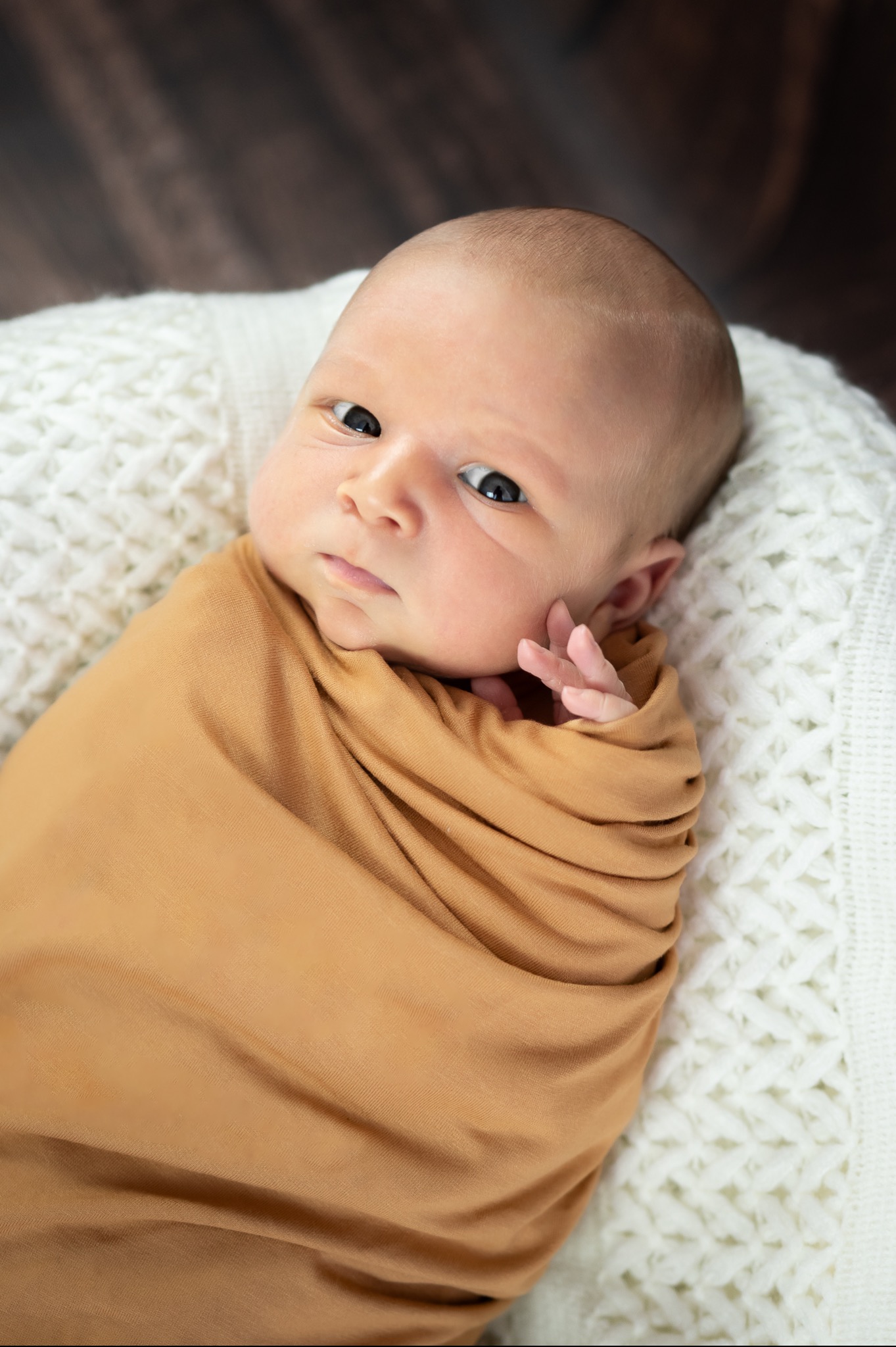 Noworodek zawinięty w musztardowy kocyk na białym dzierganym tle, z delikatnie uniesioną rączką przy policzku i skupionym spojrzeniem.