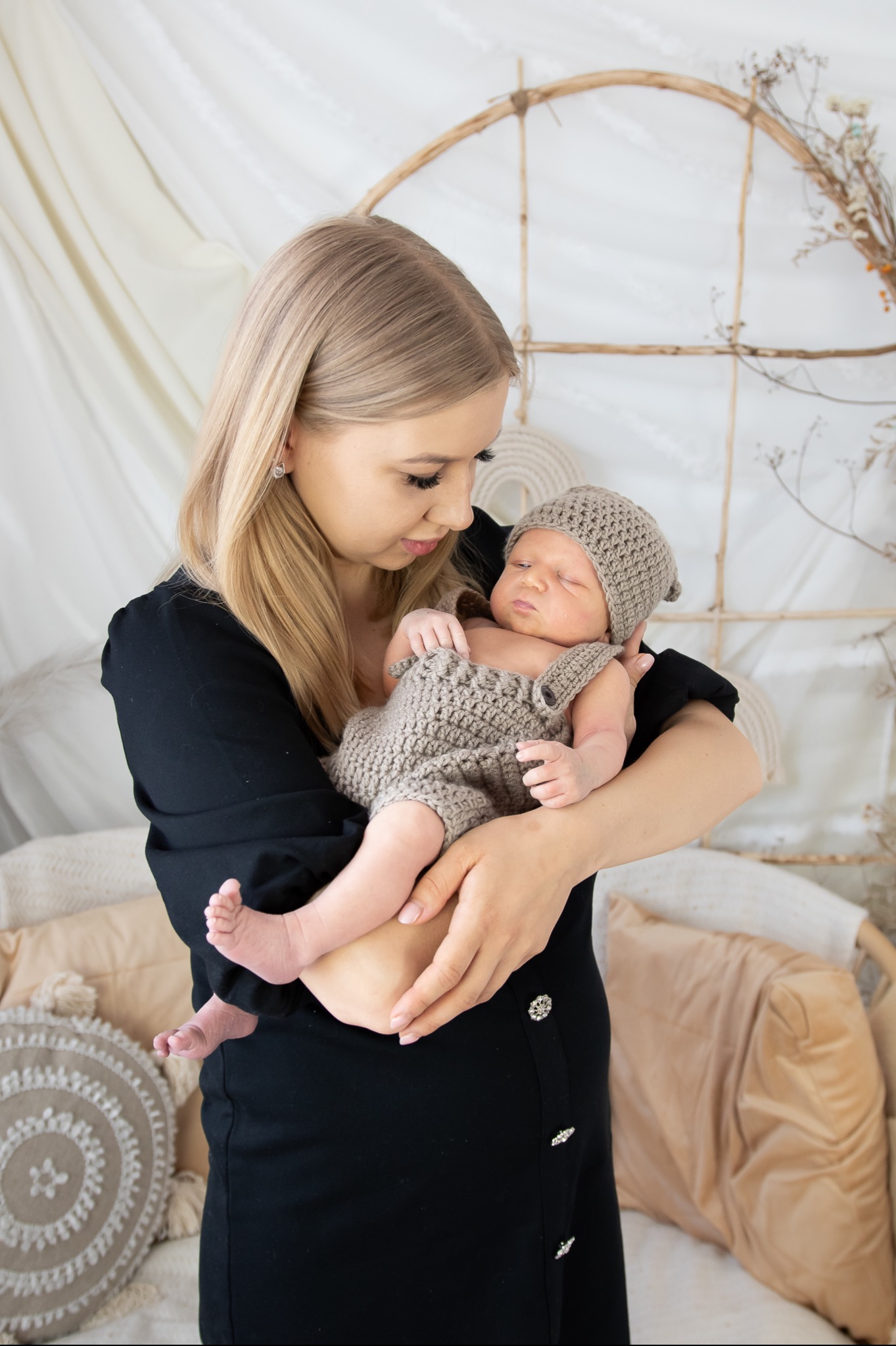 Kobieta w czarnej sukience trzyma śpiącego noworodka ubranego w dziergany kombinezon i czapeczkę na tle dekoracji z suszonych kwiatów i kremowych poduszek.